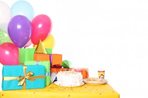 Una tavola con tovaglia gialla con sopra dei regali, dei palloncini ed una torta di compleanno